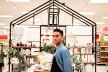 A person stands in a store aisle surrounded by various home decor items. There are small potted plants, rolled towels, and jars on the shelves. The area is lit with bright lights from the ceiling.