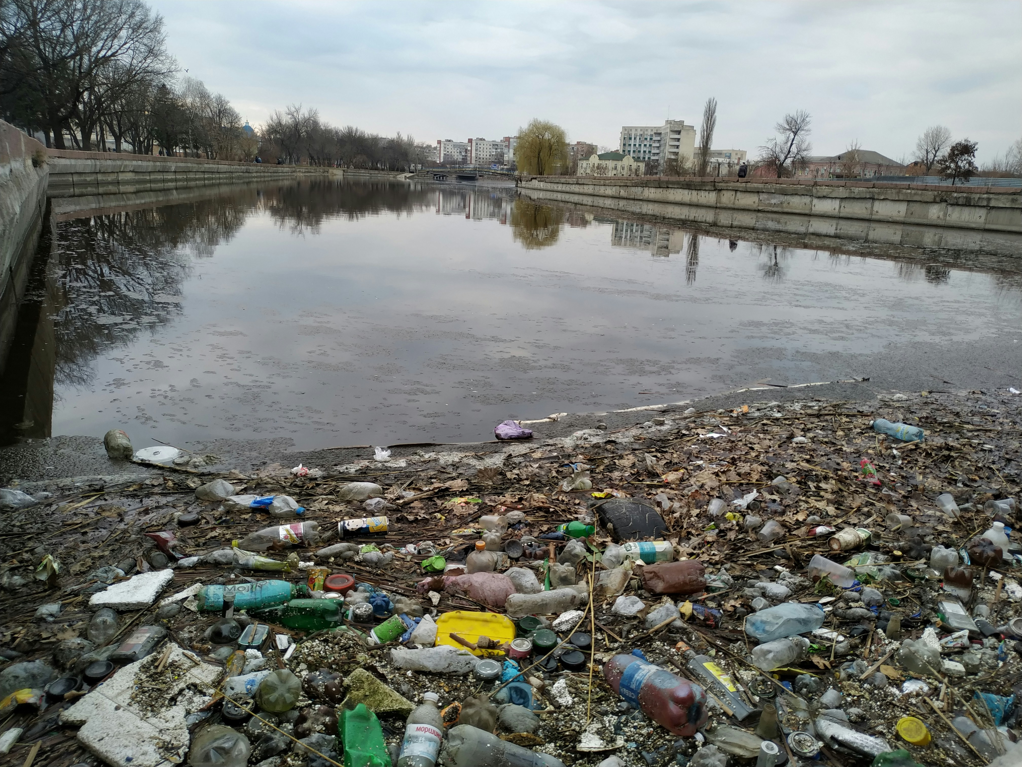 Lumë i ndotur me mbeturina plastike, ujë i turbullt, gjethe të rënë mbi sipërfaqe