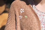 A close-up of a textured brown sweater adorned with embroidered flowers, one white with a yellow center and one small green leaf design. Part of the person's floral-patterned shirt is visible underneath the sweater.