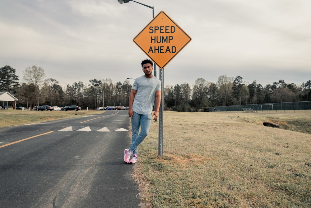 man in gray shirt leaning on speed hump ahead signage beside the road