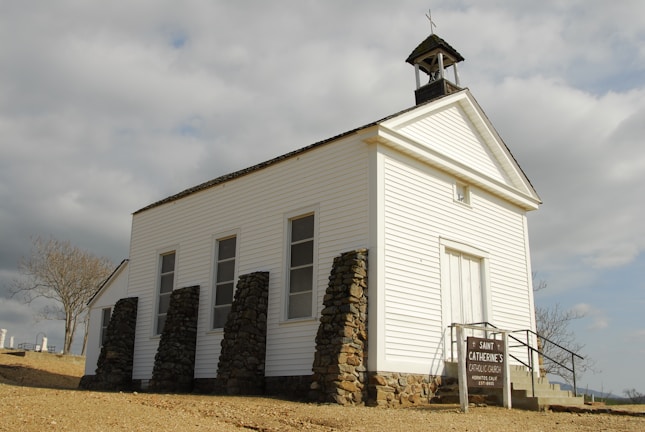 A small, white, wooden church with a triangular roof and a belfry on top, surrounded by stone buttresses. A sign in front reads 'Saint Catherine's Catholic Church'. The sky is overcast with gray clouds, and a barren tree is seen in the background. The building is situated on a slightly elevated, dirt-covered area.
