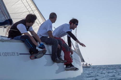 Three people are seated on the edge of a sailing boat, appearing to be engaged in an activity of concentration and teamwork. One individual, wearing a headband and sunglasses, leans forward with an outstretched arm. The boat's sails are visible, and the water is calm, with another sailboat visible in the distance.
