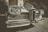A vintage car is prominently displayed in the foreground with a chrome grill and round headlights gleaming in the light. The setting seems to be outdoors, near a classic-style building partially visible in the background. The image has a retro, sepia-toned effect that emphasizes the car's classic features.