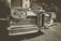 A vintage car is prominently displayed in the foreground with a chrome grill and round headlights gleaming in the light. The setting seems to be outdoors, near a classic-style building partially visible in the background. The image has a retro, sepia-toned effect that emphasizes the car's classic features.