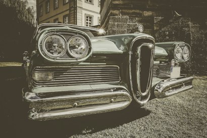 A vintage car is prominently displayed in the foreground with a chrome grill and round headlights gleaming in the light. The setting seems to be outdoors, near a classic-style building partially visible in the background. The image has a retro, sepia-toned effect that emphasizes the car's classic features.