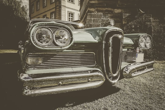 A vintage car is prominently displayed in the foreground with a chrome grill and round headlights gleaming in the light. The setting seems to be outdoors, near a classic-style building partially visible in the background. The image has a retro, sepia-toned effect that emphasizes the car's classic features.