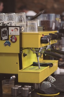 A bright yellow espresso machine adorned with various stickers and badges, including humorous coffee-themed ones. There are several clear glasses placed on top of the machine, with some mugs and metallic milk frothing pitchers on the counter nearby. The background is blurred, suggesting a cozy coffee shop environment.