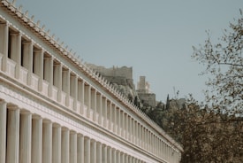 A row of classical Greek columns leads the eye toward ancient ruins partially hidden by foliage. The architecture features repetitive geometric designs and a soft, weathered appearance. In the background, a hill rises covered in trees and additional ruins, evoking a sense of history and grandeur.
