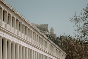 A row of classical Greek columns leads the eye toward ancient ruins partially hidden by foliage. The architecture features repetitive geometric designs and a soft, weathered appearance. In the background, a hill rises covered in trees and additional ruins, evoking a sense of history and grandeur.
