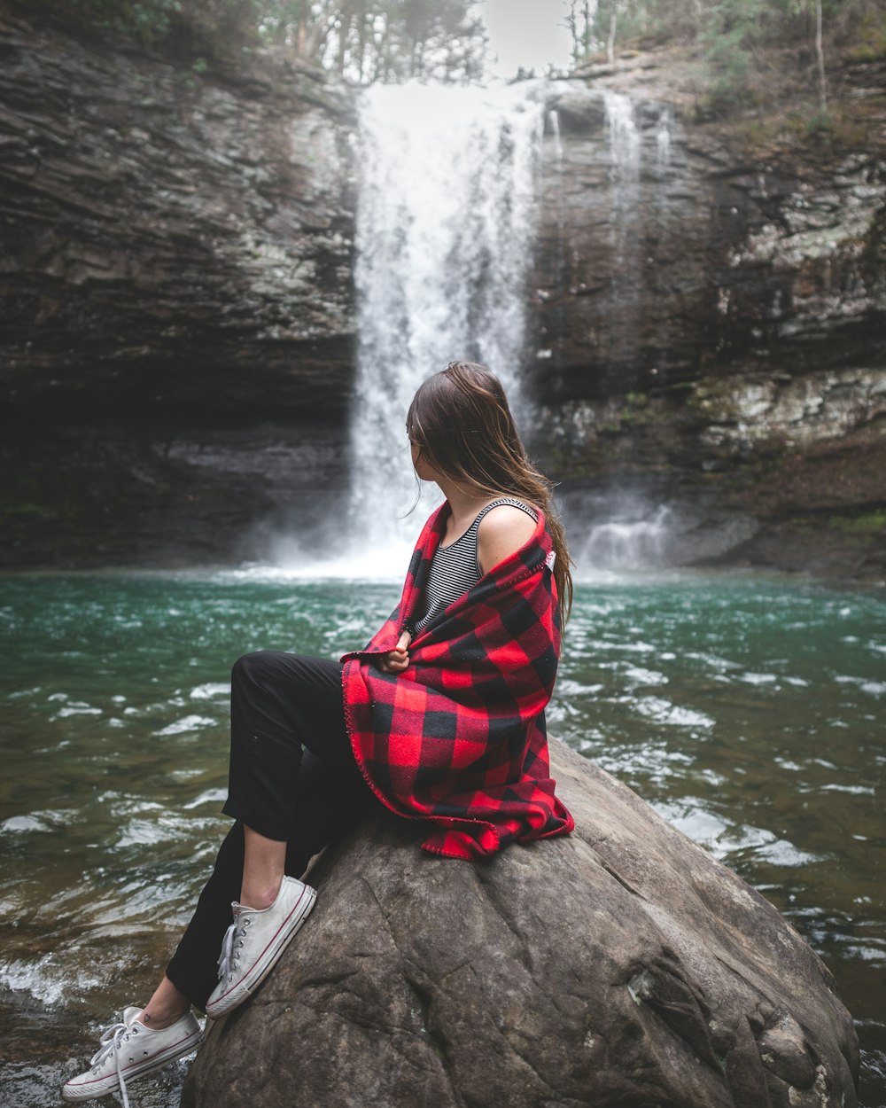 Frau in rot-schwarzem Oberteil sitzt tagsüber auf einem großen Felsen und beobachtet Wasserfälle