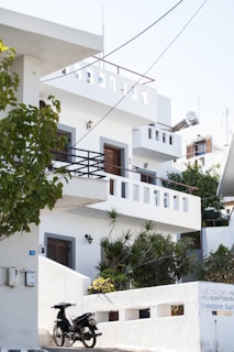 A white, multi-story building with balconies and a flat roof, situated amidst greenery. The architecture is simple and modern with white walls accented by grey edges around doors and windows. A scooter is parked by the entrance, next to a sign with visible text. The scene is lit by bright daylight, creating a clean and serene environment.