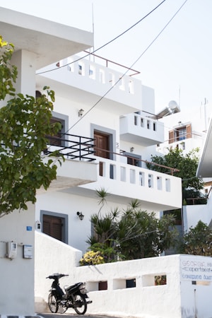 A white, multi-story building with balconies and a flat roof, situated amidst greenery. The architecture is simple and modern with white walls accented by grey edges around doors and windows. A scooter is parked by the entrance, next to a sign with visible text. The scene is lit by bright daylight, creating a clean and serene environment.