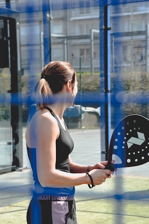 A woman holds a padel racket while standing on a court surrounded by blue fencing. She is dressed in sportswear, including a black tank top. The padel court is outdoors, with a background that includes buildings and greenery.