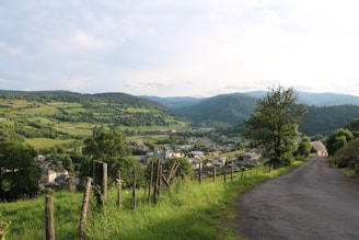 A rural landscape with a winding road leading down a hillside. Lush green fields and dense forests cover the rolling hills in the background. A small village is nestled in the valley, surrounded by scattered houses. The scene is bathed in warm, natural sunlight.