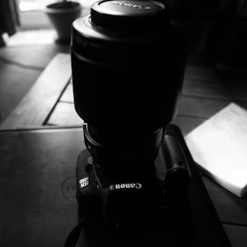A black and white photograph of a Canon EOS 600D camera placed on a table. The camera is oriented with its large lens facing upwards. Light from a nearby window casts shadows, creating a contrast between the camera and its surroundings.