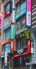 A bustling urban street scene featuring multiple storefronts adorned with vibrant signs in both English and Korean. The buildings are closely packed together, and the bright neon signs advertise various businesses such as a nail bar, Korean restaurant, eyelash salon, and karaoke lounge. The architecture is a mix of modern and older urban design.