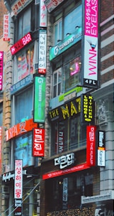 A bustling urban street scene featuring multiple storefronts adorned with vibrant signs in both English and Korean. The buildings are closely packed together, and the bright neon signs advertise various businesses such as a nail bar, Korean restaurant, eyelash salon, and karaoke lounge. The architecture is a mix of modern and older urban design.