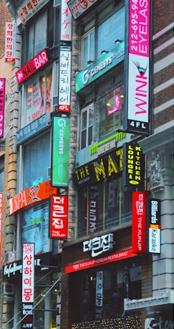 A bustling urban street scene featuring multiple storefronts adorned with vibrant signs in both English and Korean. The buildings are closely packed together, and the bright neon signs advertise various businesses such as a nail bar, Korean restaurant, eyelash salon, and karaoke lounge. The architecture is a mix of modern and older urban design.