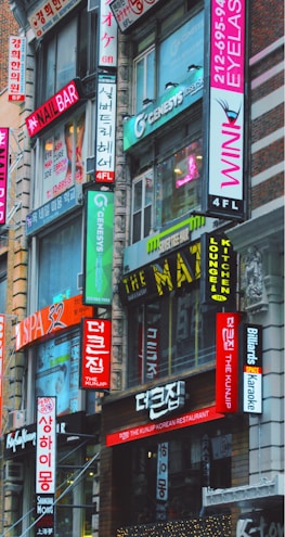 A bustling urban street scene featuring multiple storefronts adorned with vibrant signs in both English and Korean. The buildings are closely packed together, and the bright neon signs advertise various businesses such as a nail bar, Korean restaurant, eyelash salon, and karaoke lounge. The architecture is a mix of modern and older urban design.