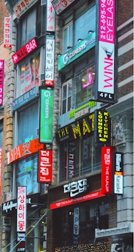 A bustling urban street scene featuring multiple storefronts adorned with vibrant signs in both English and Korean. The buildings are closely packed together, and the bright neon signs advertise various businesses such as a nail bar, Korean restaurant, eyelash salon, and karaoke lounge. The architecture is a mix of modern and older urban design.