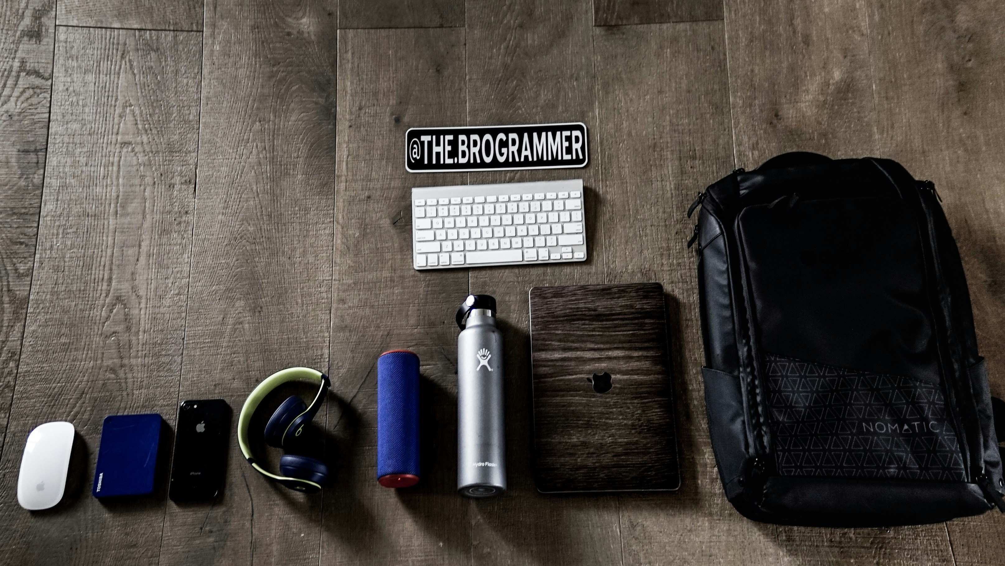 A collection of tech gadgets and personal items neatly arranged on a wooden floor. The items include a computer mouse, external hard drive, smartphone, headphones, portable speaker, water bottle, keyboard, a wooden-cased device, and a large backpack. A sticker with the text '@THE.BROGRAMMER' is placed prominently above the keyboard.