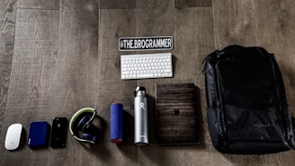 A collection of tech gadgets and personal items neatly arranged on a wooden floor. The items include a computer mouse, external hard drive, smartphone, headphones, portable speaker, water bottle, keyboard, a wooden-cased device, and a large backpack. A sticker with the text '@THE.BROGRAMMER' is placed prominently above the keyboard.