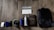 A collection of tech gadgets and personal items neatly arranged on a wooden floor. The items include a computer mouse, external hard drive, smartphone, headphones, portable speaker, water bottle, keyboard, a wooden-cased device, and a large backpack. A sticker with the text '@THE.BROGRAMMER' is placed prominently above the keyboard.