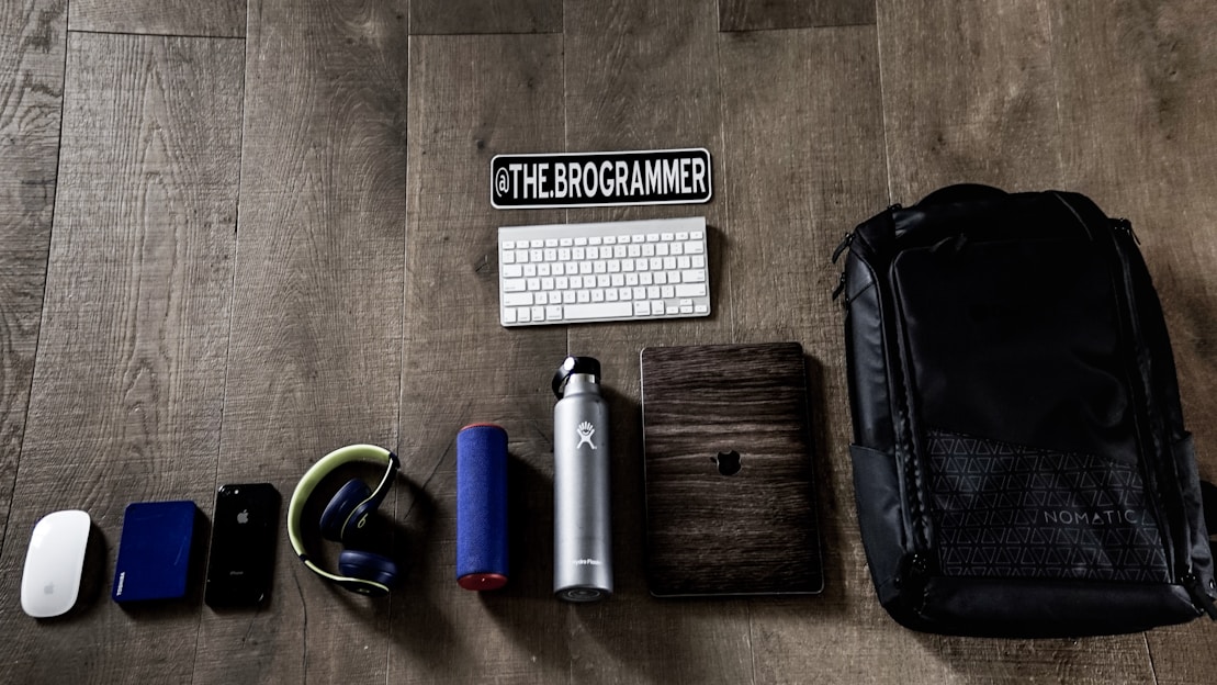 A collection of tech gadgets and personal items neatly arranged on a wooden floor. The items include a computer mouse, external hard drive, smartphone, headphones, portable speaker, water bottle, keyboard, a wooden-cased device, and a large backpack. A sticker with the text '@THE.BROGRAMMER' is placed prominently above the keyboard.