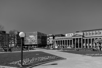 An urban plaza features a tall lamp post and manicured flower beds, surrounded by buildings with classical and modern architecture. People are walking, sitting on benches, and biking, creating a lively atmosphere.