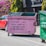 A pink dumpster with black stenciled text saying 'i thought you were special... i thought you should know' is placed on a street. It's positioned next to a red car and another green dumpster. In the background, there are lush green plants and magenta flowers against a dark building wall.
