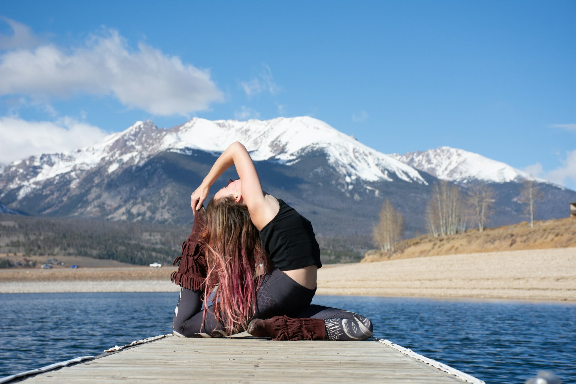 Woman on a lakeside dock with mountains behind her, arching back and reaching her arms overhead toward her back foot