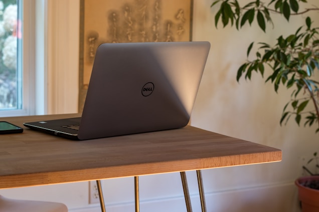A silver laptop with a logo sits open on a wooden table next to a smartphone. The table is positioned near a window, allowing natural light to illuminate the scene. A green plant is partially visible in the background, adding a touch of nature to the interior setting.