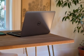A silver laptop with a logo sits open on a wooden table next to a smartphone. The table is positioned near a window, allowing natural light to illuminate the scene. A green plant is partially visible in the background, adding a touch of nature to the interior setting.