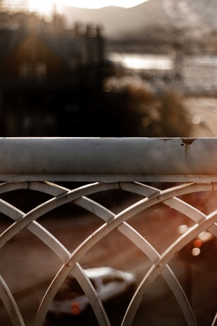 A close-up view of a decorative metal railing with a symmetrical, curved pattern is in focus. In the background, a blurred street scene with a vehicle and distant buildings is visible, bathed in warm, late-afternoon light.