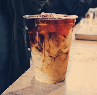A clear plastic cup filled with iced coffee, showing a dynamic swirl of cream mixing into the dark coffee. The cup sits on a slightly reflective surface, with blurred background details that suggest a casual or bustling environment.