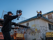 A person with a camera stabilizer films another person sitting on a rooftop. The rooftop and surrounding walls are covered with graffiti. The atmosphere appears casual and creative.