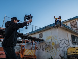 A person with a camera stabilizer films another person sitting on a rooftop. The rooftop and surrounding walls are covered with graffiti. The atmosphere appears casual and creative.