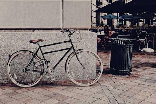 A vintage-style bicycle is leaned against a rough-textured stone wall. The bicycle has a black frame with a brown leather seat and handlebars. To the right, a metal trash can is positioned on a patterned brick pavement. In the background, there is an outdoor caf&eacute; setting with tables, chairs, and large umbrellas. Several people are sitting and chatting at the caf&eacute;.