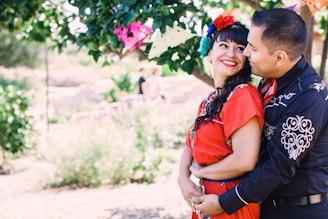 A couple stands embraced under the shade of a tree, with the woman wearing a vibrant red dress adorned with colorful embroidery and flowers in her hair. The man is dressed in a dark shirt with detailed patterns. They are both smiling warmly at each other, creating a joyful and affectionate atmosphere. The background features outdoor greenery and blurred figures in the distance, enhancing the intimate setting.