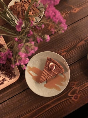 A slice of chocolate dessert topped with a pecan and cream on a light-colored plate, surrounded by artistic drizzles of caramel. The plate rests on a dark wooden table, with purple flowers partially visible in the foreground, adding a lively splash of color.
