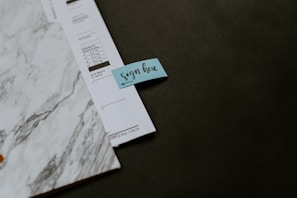 A close-up of a document with a marbled design placed on a dark surface. There is a small blue sticky note attached to the document with the words 'sign here' written on it.