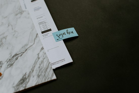 A close-up of a document with a marbled design placed on a dark surface. There is a small blue sticky note attached to the document with the words 'sign here' written on it.