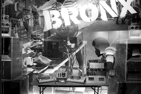 A black and white image featuring a turntable and audio equipment set up on a table. Surrounding the table are large speakers and amplifiers, with the word 'Bronx' prominently displayed in bold letters. The background shows a busy street scene with people and various musical instruments, creating a vintage urban vibe.
