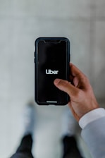 A person holds a smartphone displaying the Uber logo on the screen. The phone is oriented vertically and appears to be in use. The person is dressed in a light-colored sleeve, and the background is a neutral, blurry surface.