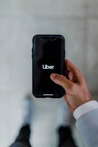 A person holds a smartphone displaying the Uber logo on the screen. The phone is oriented vertically and appears to be in use. The person is dressed in a light-colored sleeve, and the background is a neutral, blurry surface.