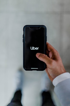 A person holds a smartphone displaying the Uber logo on the screen. The phone is oriented vertically and appears to be in use. The person is dressed in a light-colored sleeve, and the background is a neutral, blurry surface.
