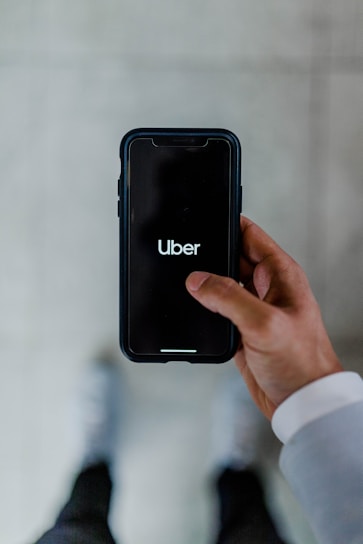 A person holds a smartphone displaying the Uber logo on the screen. The phone is oriented vertically and appears to be in use. The person is dressed in a light-colored sleeve, and the background is a neutral, blurry surface.