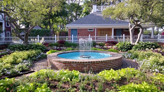 A well-maintained garden features a circular brick fountain with water gently spouting in the center, surrounded by lush greenery and colorful flowers. The background includes a charming brick house with a pitched roof, accented by white trim and a picket fence. Mature trees provide a serene, shaded atmosphere.