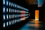 A corridor with a wall featuring multiple illuminated screens displaying abstract images in a grid pattern. The opposite wall is dark, leading to an open doorway that emits a warm orange and yellow glow. The scene has a futuristic and mysterious ambiance.