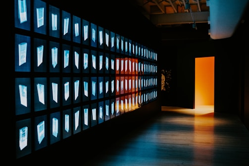 A corridor with a wall featuring multiple illuminated screens displaying abstract images in a grid pattern. The opposite wall is dark, leading to an open doorway that emits a warm orange and yellow glow. The scene has a futuristic and mysterious ambiance.